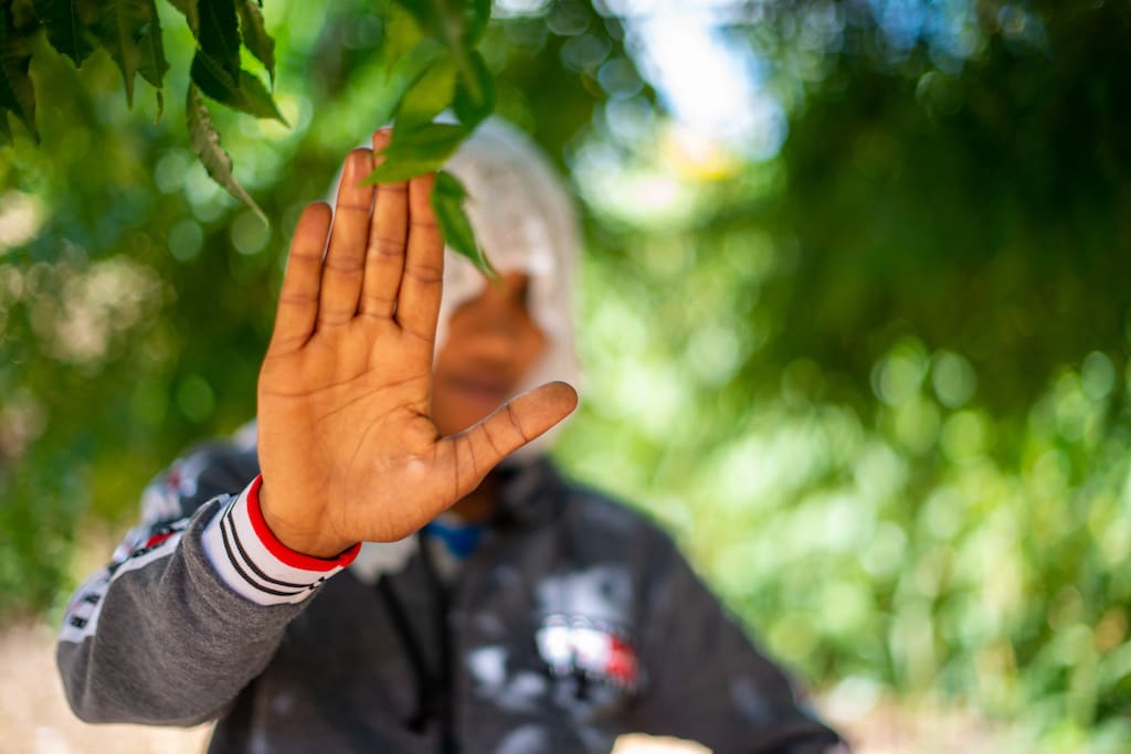 Maria is wearing a black dress and a light blue head covering. Her hand is stretched out toward the camera. The background is blurry and there are trees behind her.